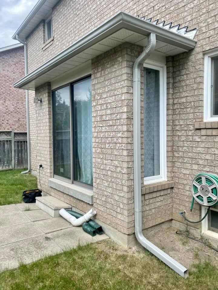 White downspout with a ground-level extension beside a brick wall.
