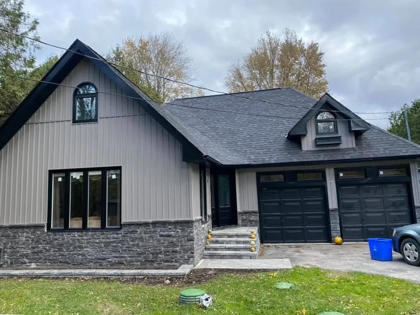 Finished home exterior with taupe board-and-batten siding, black windows, black garage doors, and a stone base.