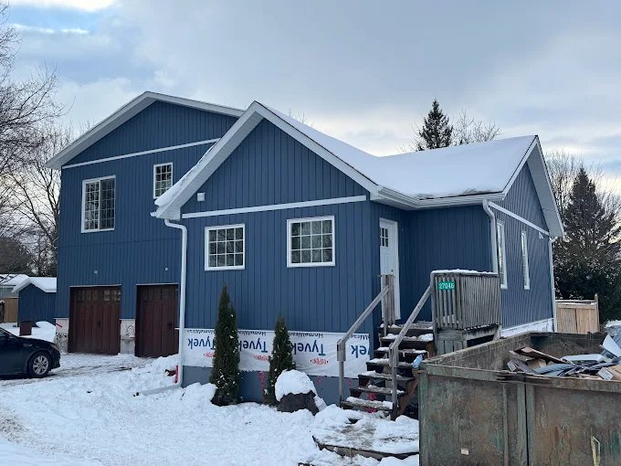 Blue board-and-batten exterior installation in winter with lower wall wrap still visible.