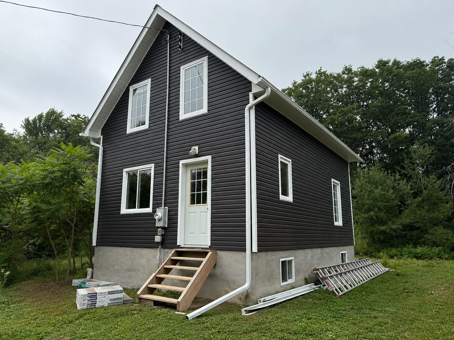 Main elevation covered in dark siding with ladders and remaining trim materials beside the house.